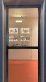 A view through a glass door shows shelves filled with various packaged goods and products. The interior is lit with warm light, and the floor is a soft orange hue. The door has inscriptions, including the phrase 'NO ENTRY' in bold letters.