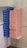 A large stack of cookie boxes is neatly arranged on two small tables. The boxes are uniformly divided into two colors: blue on the left and pink on the right. Each box has simple icon designs printed on them, and the background features a tiled wall with a light, neutral tone.