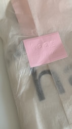 A partially open pink paper envelope features an embossed label with the text 'haag is for'. The light pink shade of the envelope contrasts subtly against the softer cream color of the paper beneath it.