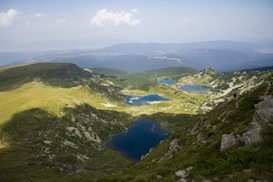 A breathtaking view of a mountainous landscape with several clear blue lakes nestled among lush green hills. The scene includes patches of sunlight and shadow cast by the clouds above. Sparse vegetation is visible on the slopes, and a few people are seen trekking along the rocky path on the right side.
