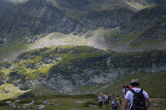A group of hikers is walking on a grassy trail at the foothills of a large, rocky mountain range. The mountain has rugged terrain with patches of greenery and areas of exposed rock. The hikers are equipped with backpacks and walking gear. The scene appears to be in a remote, natural setting.