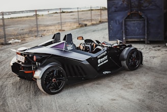 A sleek, black sports car with an open cockpit design is parked on a gravel surface near a fenced area and a large blue container. The car features branding and logos on its side, shiny black rims, and a visible license plate. The background includes sandy terrain and a distant body of water.