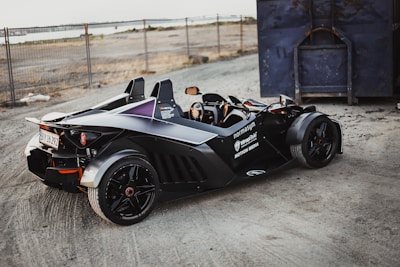 A sleek, black sports car with an open cockpit design is parked on a gravel surface near a fenced area and a large blue container. The car features branding and logos on its side, shiny black rims, and a visible license plate. The background includes sandy terrain and a distant body of water.