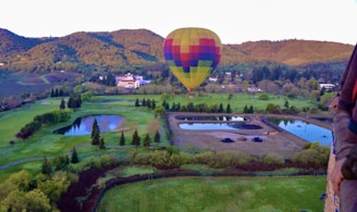 A hot air balloon with a patchwork of colorful squares floating above a lush green landscape. The area below includes manicured gardens, several bodies of water, and a number of deciduous trees. There are rolling green hills in the background, and a large building can be seen near the center of the image. The scene is framed by a hot air balloon basket.