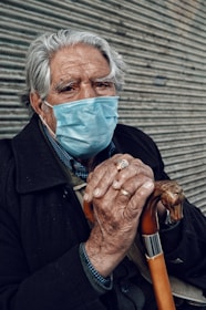 An elderly person with gray hair is wearing a blue surgical mask and a dark coat, holding a walking cane with both hands. The background features a corrugated metal surface.