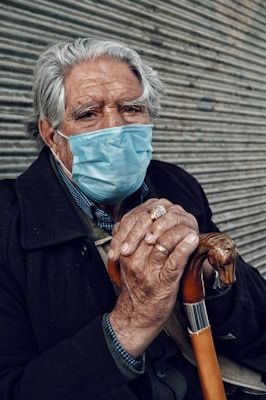An elderly person with gray hair is wearing a blue surgical mask and a dark coat, holding a walking cane with both hands. The background features a corrugated metal surface.