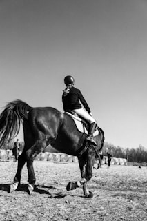 A person wearing a helmet rides a horse on a dirt field. The rider is dressed in equestrian attire. In the background, there are hay bales and a few people standing or walking.