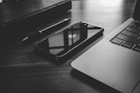 A black and white photo displays a sleek smartphone with a visible logo placed next to a laptop. A pen and a closed notebook are also present on the desk. The reflective surface of the phone shows a window pattern.