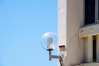 A modern, clear glass light fixture is mounted on an exterior wall of a building. The wall is painted in a light, neutral color with some visible weathering. The sky is clear and bright blue, suggesting a sunny day.