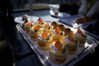 A tray of beautifully arranged desserts featuring small, round cakes topped with vibrant orange slices and garnishes, possibly mint. The cakes are neatly organized and displayed on a white rectangular plate, with soft lighting creating a warm, inviting atmosphere.
