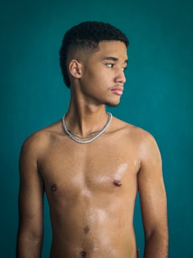 A young man stands against a teal background, looking to his right. He has a short haircut and wears a silver chain necklace. His upper body glistens as if wet or oiled, displaying a smooth, toned physique.