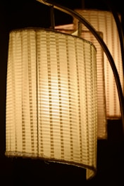 Warm-toned lampshades with vertical striped patterns are illuminated in a soft, golden light against a dark background. The woven texture suggests a handmade or artisanal design.