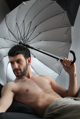 A shirtless man with short dark hair is reclining on a black surface while holding a large, open silver umbrella over his shoulder. He appears to be indoors, and the background is a plain grey wall.
