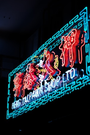 A vibrant neon sign displaying the text 'Heng Tai Pawn Shop Ltd' in English with decorative Asian characters above. The sign is illuminated with bright colors against a dark background.