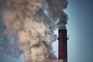 A large industrial smokestack emits a substantial amount of thick, dark smoke into the air, against a backdrop of a clear sky.