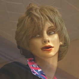 A close-up view of a mannequin head with realistic facial features and short, wavy brown hair. The mannequin wears a shirt with a colorful collar featuring red, blue, and white patterns. The lighting creates a soft, warm effect on the mannequin's face.