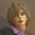 A close-up view of a mannequin head with realistic facial features and short, wavy brown hair. The mannequin wears a shirt with a colorful collar featuring red, blue, and white patterns. The lighting creates a soft, warm effect on the mannequin's face.
