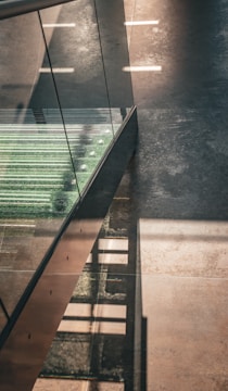 A modern staircase featuring glass sides and metallic steps with a greenish texture, casting geometric shadows on a polished floor. The lighting creates strong contrasts between the illuminated surfaces and areas in shadow, enhancing the architectural elements.