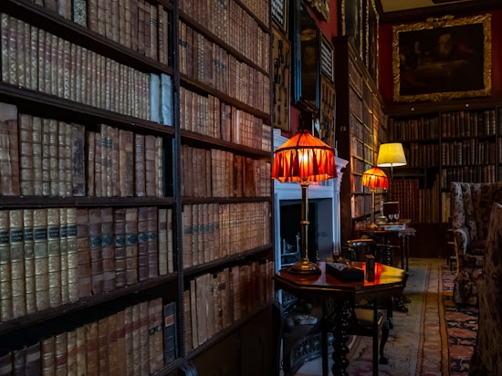 A cozy library filled with numerous old books on wooden shelves. A warm atmosphere is created by the soft glow of several ornate lamps with red and amber shades. The room features elegant furniture and detailed carpeting, adding to the classic ambiance. There are paintings on the walls, and tables arranged with books and decorative items.