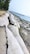 Large sandbags line a shoreline, serving as a barrier against the sea. The bags appear weathered, and the sand around them is wet and compact. A line of trees stretches along the edge, creating a natural border between the sandy area and the bags.