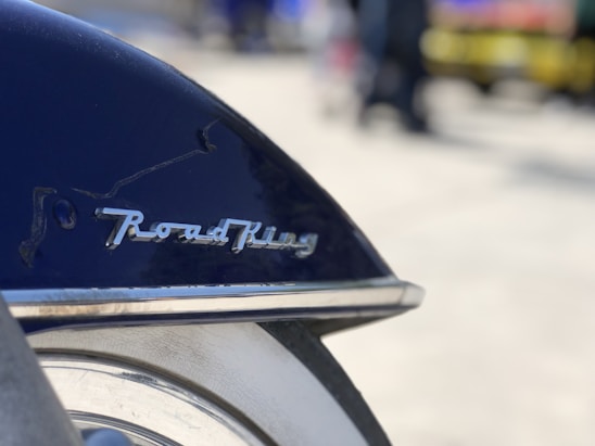 A close-up view of a motorcycle fender with the logo 'Road King' prominently displayed. The fender is dark blue, and the background is softly blurred, creating a bokeh effect.