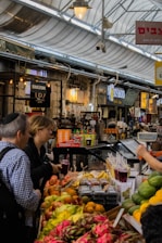 A bustling market scene featuring a variety of colorful fruits on display, including dragon fruits, oranges, and bananas. People are interacting and purchasing items at the stall lined with juices in plastic cups. The market is covered with a transparent roof, and several signs in different languages are visible.