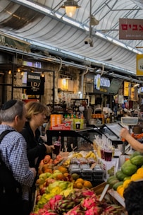 A bustling market scene featuring a variety of colorful fruits on display, including dragon fruits, oranges, and bananas. People are interacting and purchasing items at the stall lined with juices in plastic cups. The market is covered with a transparent roof, and several signs in different languages are visible.