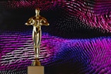 A golden statue resembling an award is centrally placed against a dynamic background of swirling patterns in shades of pink, purple, and blue. The statue stands on a base with the phrase 'DO WHATEVER IT TAKES' engraved on it.