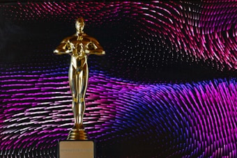 A golden statue resembling an award is centrally placed against a dynamic background of swirling patterns in shades of pink, purple, and blue. The statue stands on a base with the phrase 'DO WHATEVER IT TAKES' engraved on it.