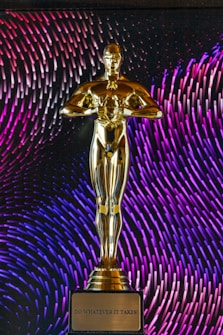 A golden statuette stands prominently on a base with an inscription that reads 'Do Whatever It Takes.' In the background, vibrant patterns of purple and blue lines create a dynamic and colorful backdrop.