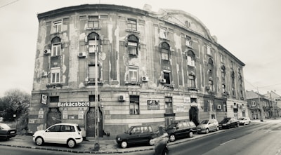 A sepia-toned photograph of a large, aged building on a street corner. The structure has worn and peeling paint, with multiple windows and small balconies. Several cars are parked along the street, and a few signs are visible, including one labeled 'Barkácsbolt'. A person is captured in motion, walking by.