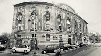 A sepia-toned photograph of a large, aged building on a street corner. The structure has worn and peeling paint, with multiple windows and small balconies. Several cars are parked along the street, and a few signs are visible, including one labeled 'Barkácsbolt'. A person is captured in motion, walking by.