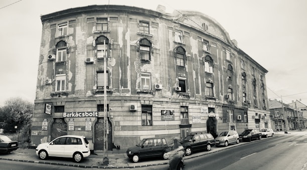 A sepia-toned photograph of a large, aged building on a street corner. The structure has worn and peeling paint, with multiple windows and small balconies. Several cars are parked along the street, and a few signs are visible, including one labeled 'Barkácsbolt'. A person is captured in motion, walking by.
