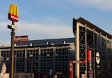 A commercial area with multiple restaurant signs visible, including McDonald's, Roadhouse Restaurant, and Mercio Sofà. The McDonald's sign is prominently displayed on a pole in the foreground. The building in the background has a modern glass facade and signage indicating the presence of drive-through services.