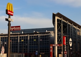 A commercial area with multiple restaurant signs visible, including McDonald's, Roadhouse Restaurant, and Mercio Sofà. The McDonald's sign is prominently displayed on a pole in the foreground. The building in the background has a modern glass facade and signage indicating the presence of drive-through services.