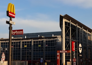 A commercial area with multiple restaurant signs visible, including McDonald's, Roadhouse Restaurant, and Mercio Sofà. The McDonald's sign is prominently displayed on a pole in the foreground. The building in the background has a modern glass facade and signage indicating the presence of drive-through services.