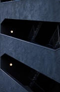 The image features a modern architectural structure with staggered, open corridors. The walls are dark, creating a stark contrast with the light coming from spherical wall lamps inside. The multi-level design is geometric, highlighting the straight lines of railings and stairs.