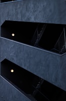 The image features a modern architectural structure with staggered, open corridors. The walls are dark, creating a stark contrast with the light coming from spherical wall lamps inside. The multi-level design is geometric, highlighting the straight lines of railings and stairs.