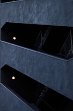 The image features a modern architectural structure with staggered, open corridors. The walls are dark, creating a stark contrast with the light coming from spherical wall lamps inside. The multi-level design is geometric, highlighting the straight lines of railings and stairs.