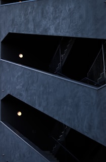The image features a modern architectural structure with staggered, open corridors. The walls are dark, creating a stark contrast with the light coming from spherical wall lamps inside. The multi-level design is geometric, highlighting the straight lines of railings and stairs.
