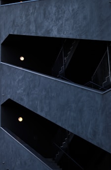 The image features a modern architectural structure with staggered, open corridors. The walls are dark, creating a stark contrast with the light coming from spherical wall lamps inside. The multi-level design is geometric, highlighting the straight lines of railings and stairs.