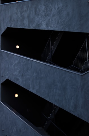 The image features a modern architectural structure with staggered, open corridors. The walls are dark, creating a stark contrast with the light coming from spherical wall lamps inside. The multi-level design is geometric, highlighting the straight lines of railings and stairs.