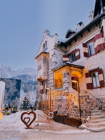 A picturesque stone building with rustic charm is decorated with festive string lights, creating a cozy and warm ambiance. Snow blankets the surrounding area, enhancing the wintery feel. A heart-shaped sculpture adds a romantic touch in the foreground, while mountains create a scenic backdrop.