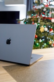 A gray laptop with an apple logo is placed on a wooden table, with a decorated Christmas tree blurred in the background. The tree is adorned with colorful ornaments, including red, white, and gold baubles. The setting suggests a cozy indoor environment with subtle lighting.