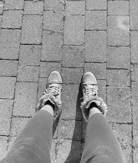 A pair of legs in leggings is visible, wearing sneakers with fur trim, standing on a paved brick pathway. The image is in black and white.
