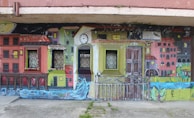A vibrant mural depicting a colorful urban scene with houses painted in various bright shades. The mural incorporates real windows into the artwork, blending seamlessly with the painted windows of the buildings. There is a clock tower at the center, and a flowing river at the bottom of the mural adds a dynamic element to the scene.
