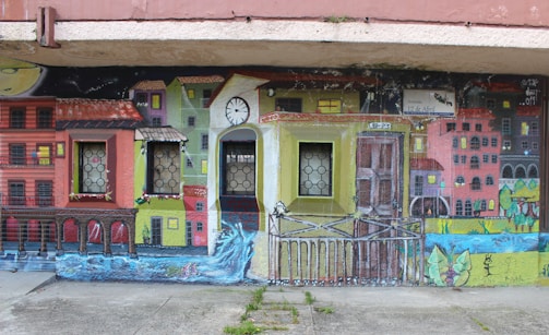 A vibrant mural depicting a colorful urban scene with houses painted in various bright shades. The mural incorporates real windows into the artwork, blending seamlessly with the painted windows of the buildings. There is a clock tower at the center, and a flowing river at the bottom of the mural adds a dynamic element to the scene.
