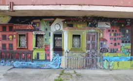 A vibrant mural depicting a colorful urban scene with houses painted in various bright shades. The mural incorporates real windows into the artwork, blending seamlessly with the painted windows of the buildings. There is a clock tower at the center, and a flowing river at the bottom of the mural adds a dynamic element to the scene.