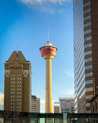 A tall observation tower stands prominently between modern skyscrapers, with a clear blue sky in the background. The tower has a unique red and beige design, and a hotel building is visible to the left.