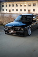 A black vintage BMW car is parked on a concrete surface with a large, beige industrial building in the background. The car is well-maintained, reflecting light on its polished exterior. The scene is set outdoors, with some dry grass and plants visible along the edge of the pavement.